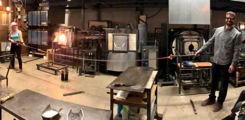 Two glassblowers in a workshop, one shaping molten glass on a rod while the other assists, surrounded by tools and equipment.