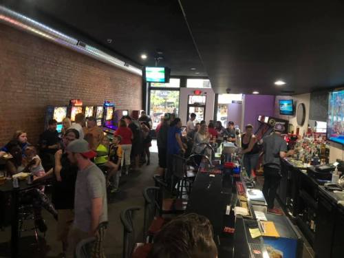 A lively bar scene with patrons enjoying drinks, arcade games, and a bartender serving at the counter.