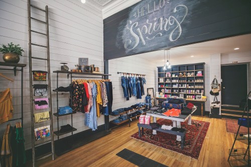 A cozy retail space featuring clothing racks, a table with merchandise, and a chalkboard welcoming spring.