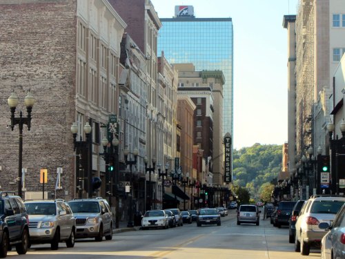 A city street lined with historic buildings and modern skyscrapers, with cars parked along the road and green hills in the background.