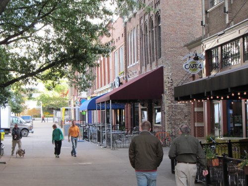 A tree-lined street with people walking, shops, and outdoor seating at a café.