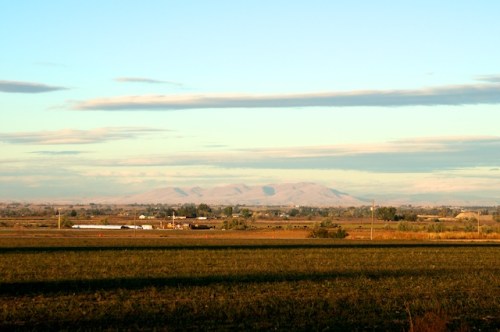 A scenic view of rolling fields with distant mountains under a clear blue sky and soft clouds.
