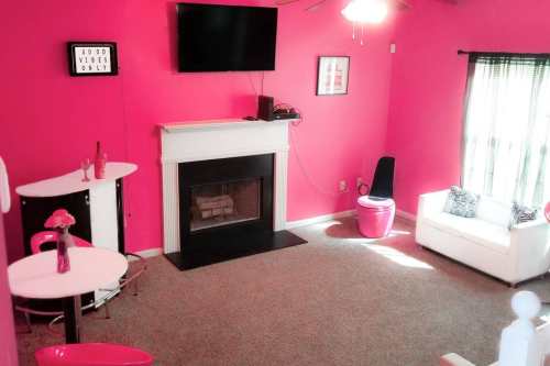 Bright pink living room with a fireplace, TV, white sofa, and bar table. Decor includes a "Good Vibes Only" sign.