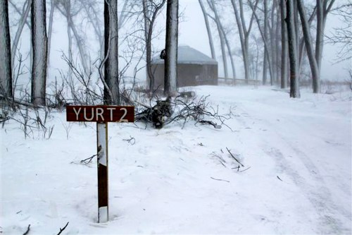 A snowy path leads to a yurt, with a sign reading "YURT2" amidst bare trees in a winter landscape.