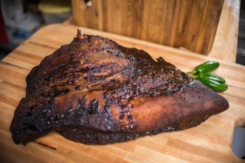 A large, smoked brisket resting on a wooden cutting board, garnished with fresh jalapeños.