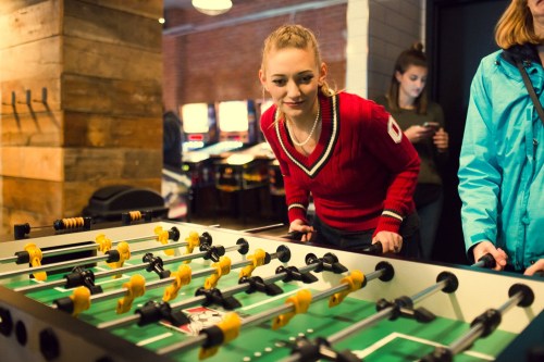 A woman in a red sweater leans over a foosball table, focused on the game, while another person stands nearby.