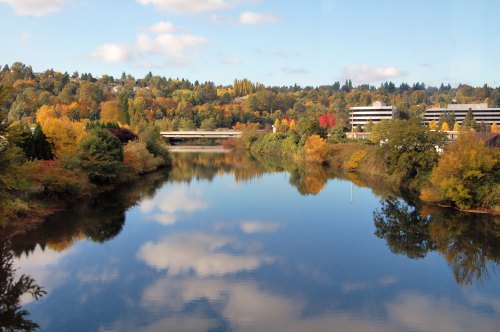 A serene river reflects colorful autumn trees and buildings under a partly cloudy sky.