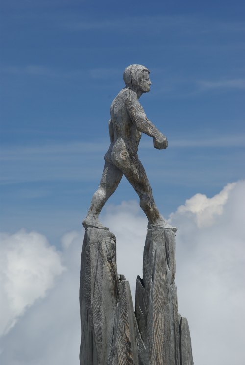 A wooden sculpture of a figure walking atop a rocky peak, set against a blue sky with clouds.