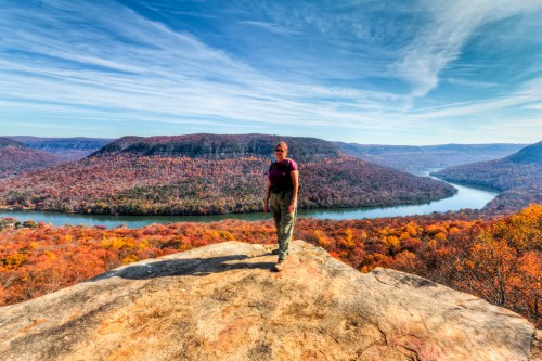 A person stands on a rocky outcrop overlooking a river and vibrant autumn foliage in a scenic landscape.