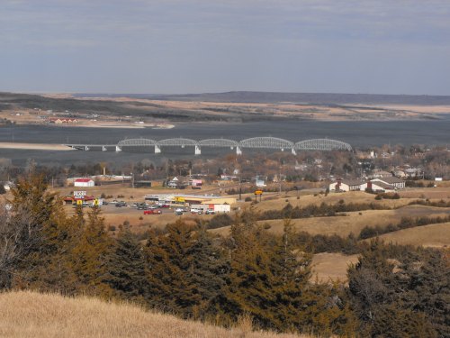 A scenic view of a bridge over a river, with a small town and rolling hills in the background.