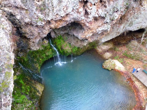 A serene waterfall cascades into a clear pool surrounded by rocky cliffs and lush greenery.