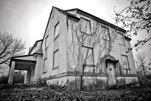 Abandoned two-story house overgrown with vines, set against a cloudy sky in black and white.