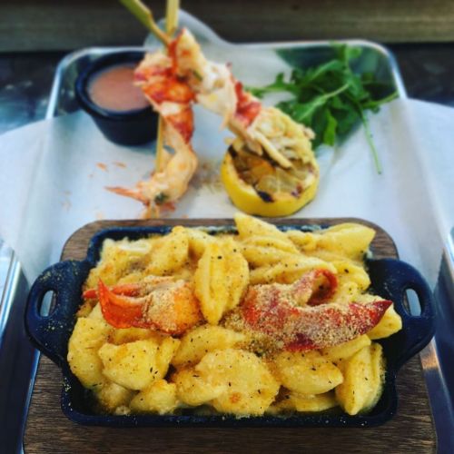 A serving of creamy lobster mac and cheese with a lobster claw, served on a wooden tray with a side of sauce.