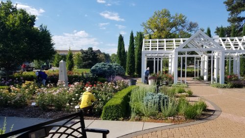 Two people tending to a colorful flower garden near a white gazebo on a sunny day.