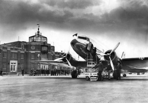 A vintage airplane is being serviced at an airport with a historic terminal building in the background.