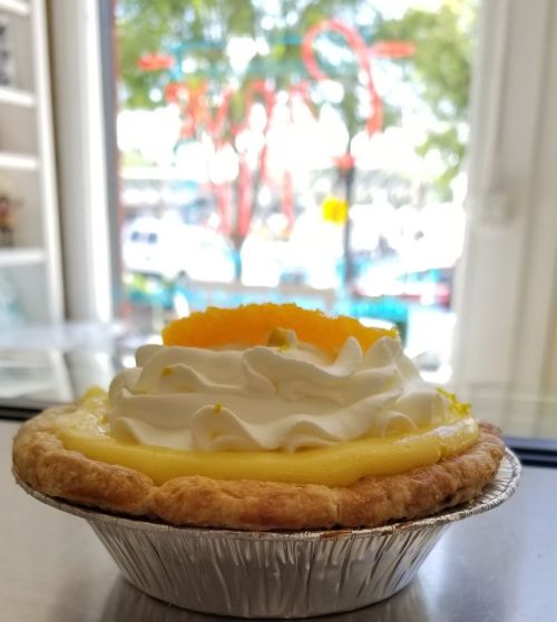 A close-up of a lemon meringue pie with whipped cream and a slice of lemon, set against a blurred window view.