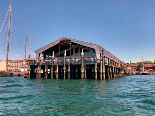 A waterfront restaurant on stilts, bustling with diners, surrounded by boats and clear blue water under a sunny sky.
