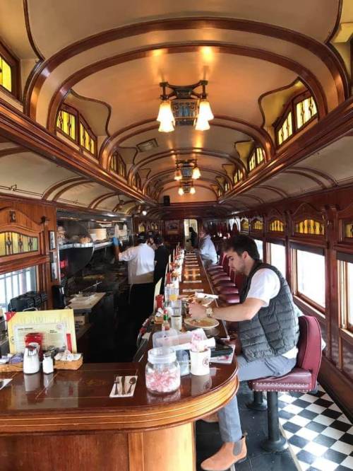 Interior of a vintage diner with a long counter, patrons seated, and staff preparing food in a cozy atmosphere.