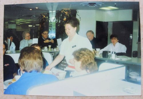 A waitress serves food in a busy diner, with customers seated at tables in the background.