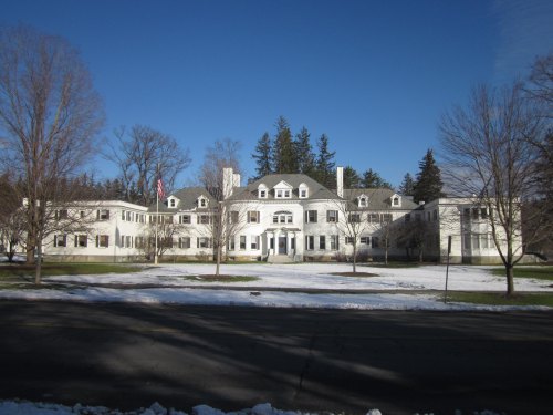 A large white building with a symmetrical facade, surrounded by trees and patches of snow under a clear blue sky.
