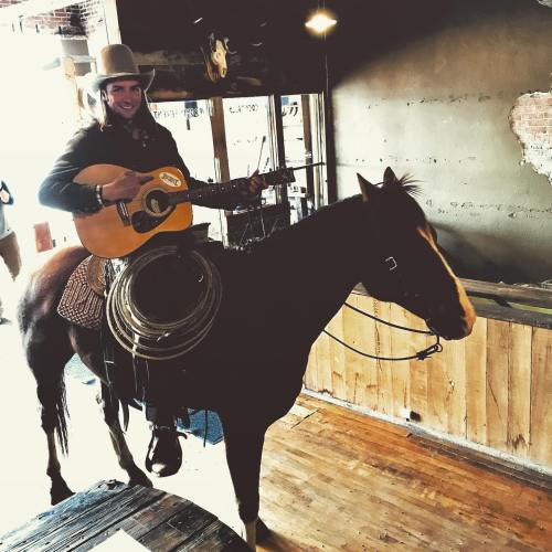 A person in a cowboy hat plays guitar while sitting on a horse inside a rustic venue.