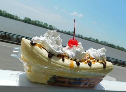 A banana split topped with whipped cream, nuts, chocolate sauce, and a cherry, with a river in the background.