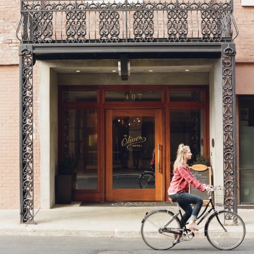 A woman rides a bicycle past the entrance of the Oliver Hotel, featuring ornate ironwork and wooden doors.
