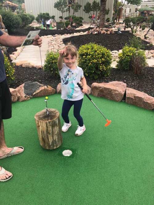A young girl in a blue shirt poses with a golf club at a mini-golf course, smiling and pointing excitedly.