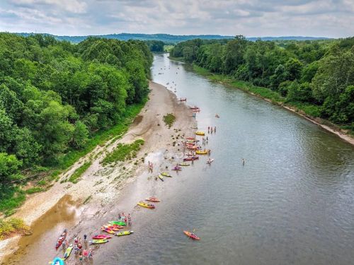 Aerial view of a river with kayakers and people on the shore, surrounded by lush green trees and a cloudy sky.