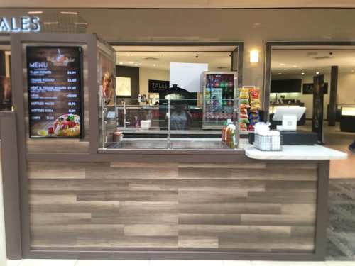 A food kiosk in a mall with a menu display, snacks, and a cash register, set against a modern interior backdrop.