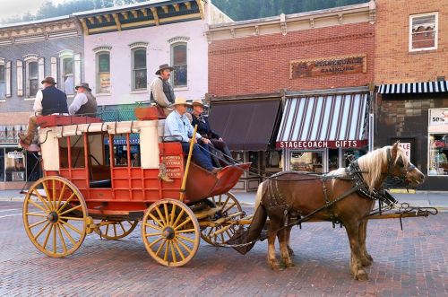 A horse-drawn stagecoach with passengers rides down a historic street lined with shops and buildings.