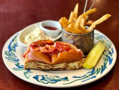 A lobster roll on a plate with fries, coleslaw, and a pickle on the side.