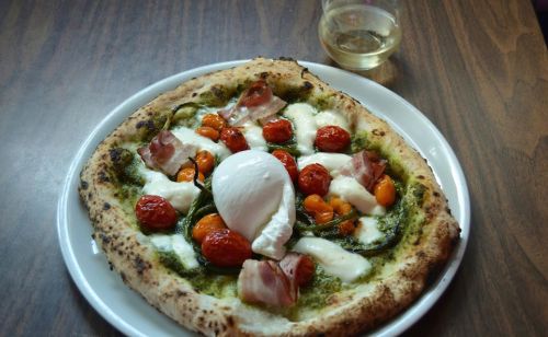 A gourmet pizza topped with cherry tomatoes, mozzarella, basil pesto, and a poached egg, served with a glass of white wine.