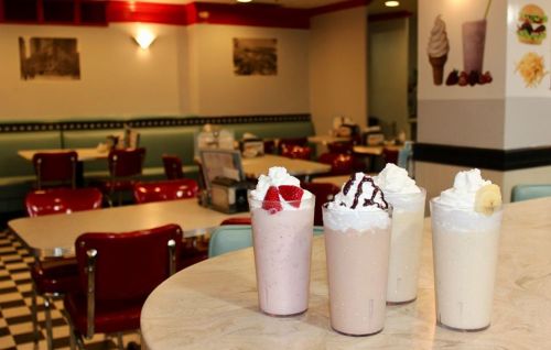 Four milkshakes topped with whipped cream and fruit sit on a table in a retro diner with red chairs and checkered floor.