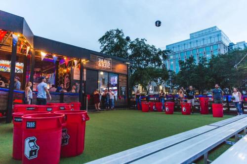 A lively outdoor venue with people playing games, colorful banners, and large red containers on a green turf.