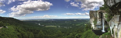 A panoramic view of a lush green valley with a waterfall cascading from a rocky cliff under a blue sky with clouds.
