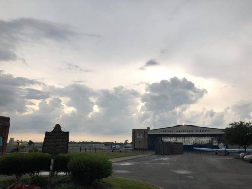 Cloudy sky over an airport hangar with "Central American Airways" sign, and a historical marker in the foreground.