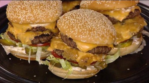 A close-up of stacked cheeseburgers with sesame seed buns, lettuce, tomato, pickles, and melted cheese.