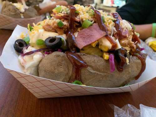 A loaded baked potato topped with cheese, barbecue sauce, olives, corn, and green onions in a paper tray.