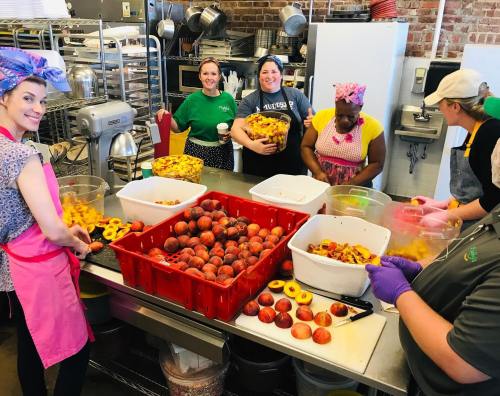 A group of women in a kitchen preparing fruits, with peaches and other produce in large containers and bowls.