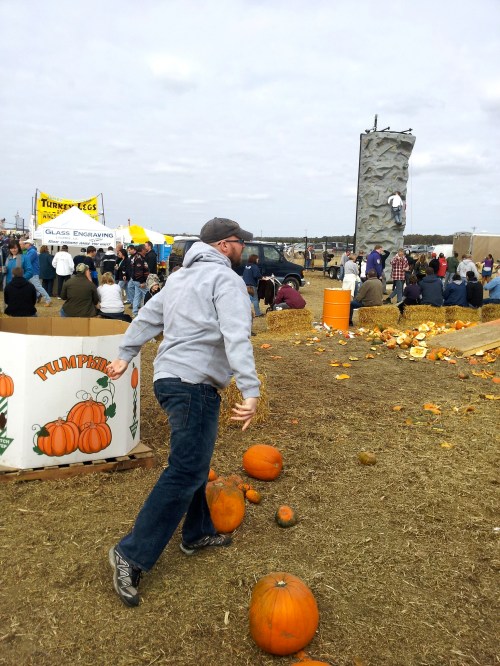 A man in a gray hoodie walks past pumpkins at a fall festival with a rock climbing wall and crowd in the background.
