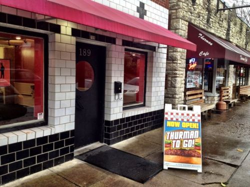 Exterior of a restaurant with a red awning and a sign reading "NOW OPEN! THURMAN TO GO!" on the sidewalk.