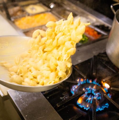 A chef pouring cooked pasta from a pan onto a stove with a blue flame, surrounded by various ingredients.