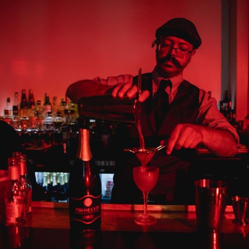 A bartender with a mustache pours a cocktail into a glass, surrounded by bottles in a dimly lit, red-tinted bar.