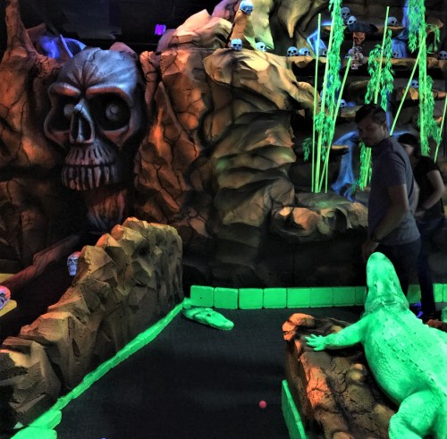 A glow-in-the-dark mini-golf course featuring a skull cave, green alligator, and vibrant decorations.