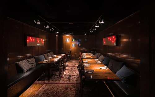 A dimly lit restaurant interior with wooden walls, long tables, and plush seating, featuring artwork on the walls.
