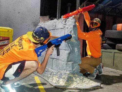 Two players in bright orange shirts aim toy blasters from behind a concrete barrier in a playful shooting arena.
