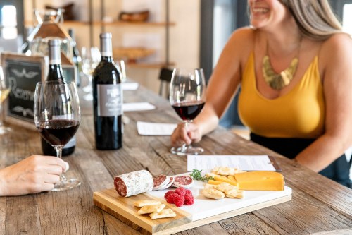 A wooden platter with cheese, salami, and raspberries, alongside two glasses of red wine and a smiling woman in a yellow top.