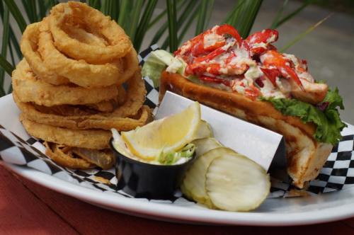A plate featuring a lobster roll with lettuce, onion rings, pickles, and lemon wedges on the side.
