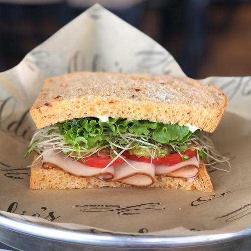 A close-up of a sandwich with turkey, lettuce, tomato, sprouts, and mayonnaise on whole grain bread, served on parchment paper.